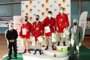 Keturi vaikinai, vilkintys raudonos spalvos kimono bei medicinines veido kaukes stovi ant apdovanojimų pakylos, iš šonų stovi du vyrai vilkintys kostiumus 
