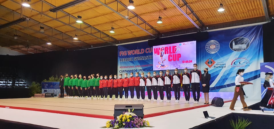 Sporto salėje stovi trys grupės sportininkų - vieni vilkintys žalius džemperius, kiti- raudonus ir treti - juodus