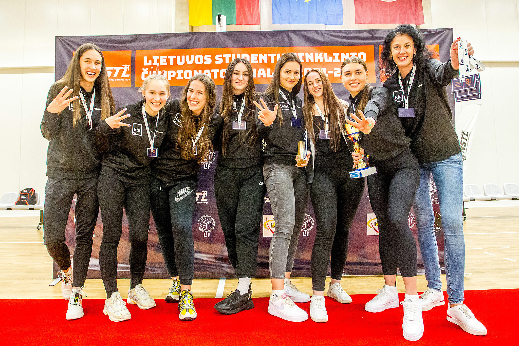 Septynios studentės tinklininkės stovi greta trenerės juodais plaukais