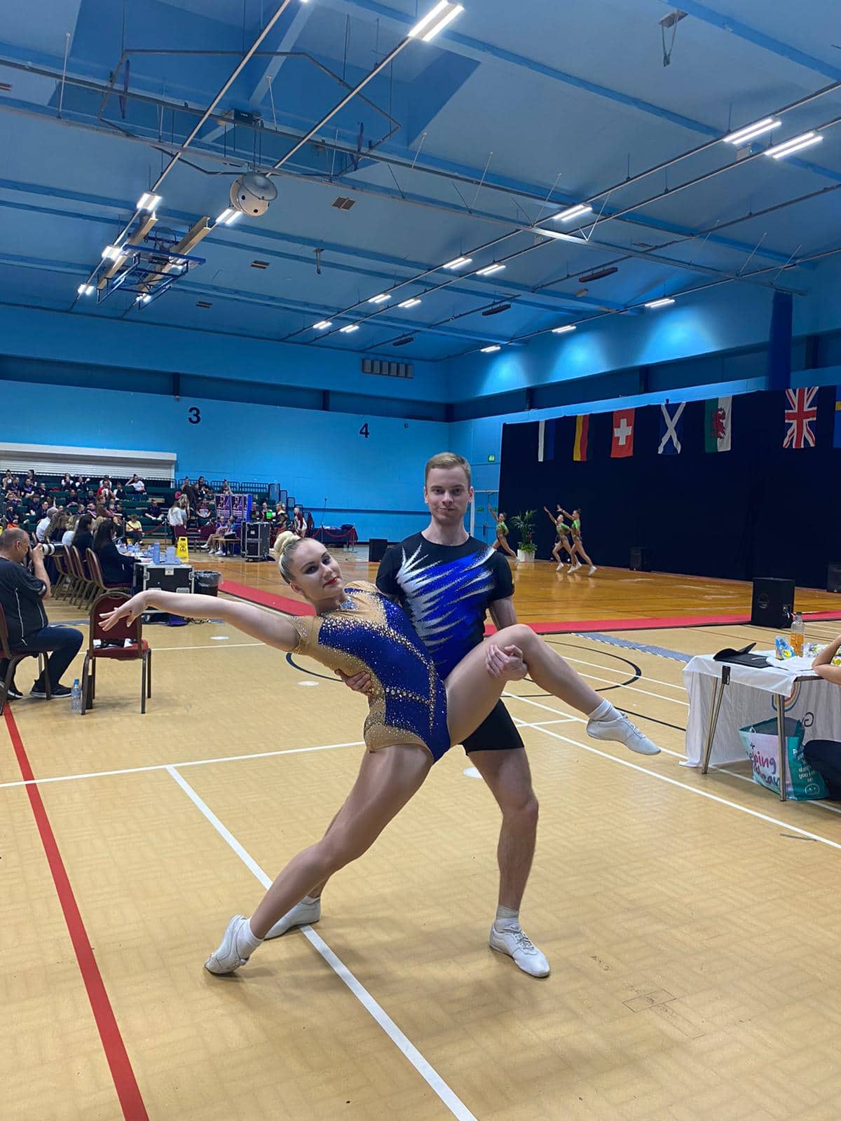 Gimnastė ir gimnastas, dėvintys mėlynos spalvos triko atlieka šokių judesį sporto salėje