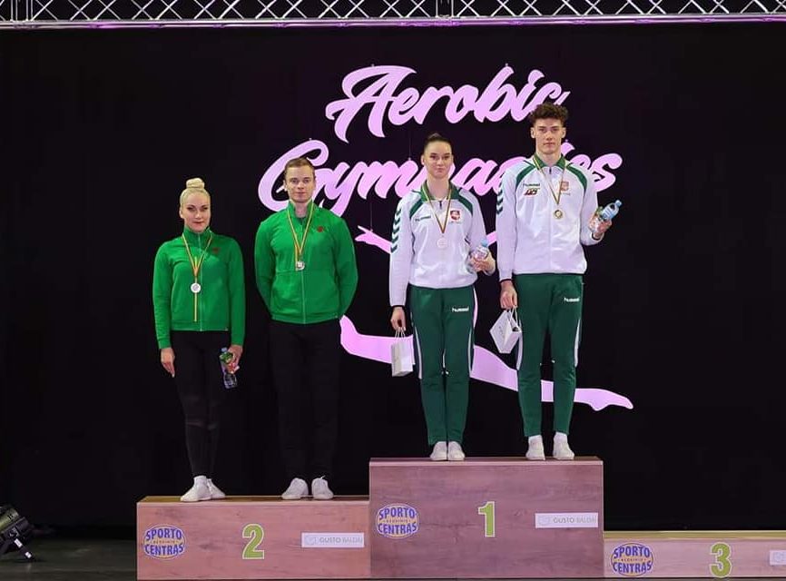 Ant apdovanojimų pakylos stovi pirmosios vietos nugalėtojai - vaikinas ir mergina dėvintys baltai žalius sportinius kostiumus, bei antros vietos nugalėtojai dėvintys žalios ir juodos spalvos sportinius kostiumus