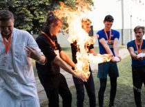 Penki vyrai laiko ištiesę rankas į priekį, vieno vyro rankose dega ugnis
