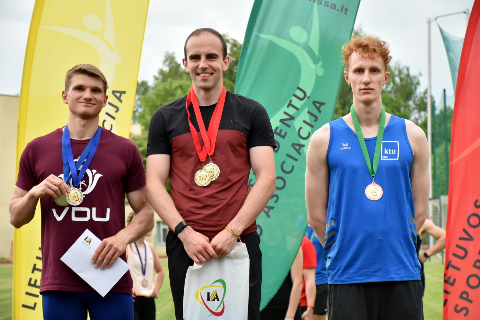Trys vaikinai stovi Lietuvos vėliavos fone, su užkabintais ant kaklo medaliais