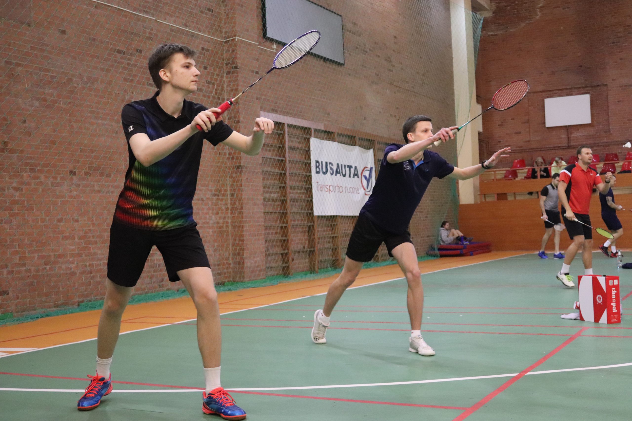 Du vaikinai studentai vilkintys juoda sportine apranga, žaidžia badmintoną badmintono aikštelėje.