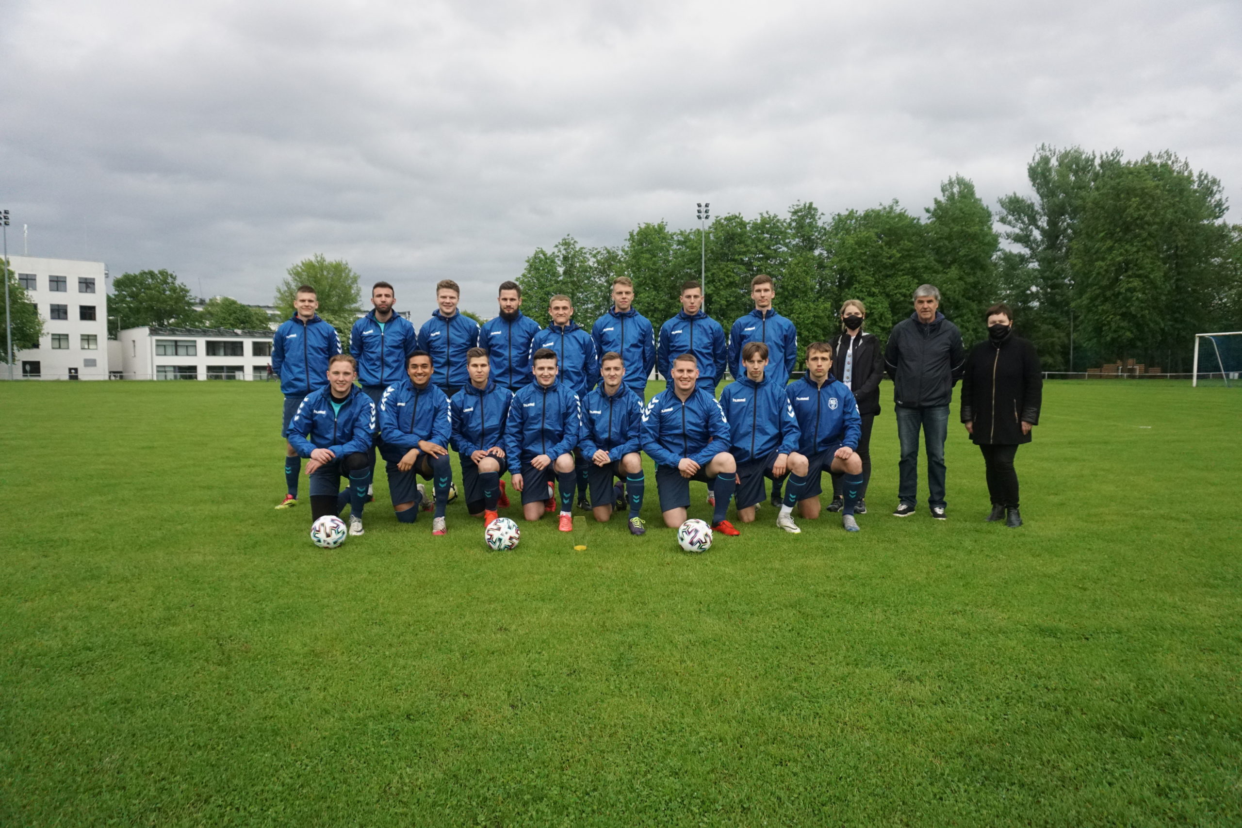 Šešiolika futbolininkų mėlynos spalvos apranga bei dvi moterys ir vyras, vilkintys juodai, stovi futbolo aikštėje