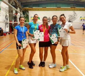 Penkios badmintono žaidėjos moterys stovi greta viena kitos ir laiko padėkas