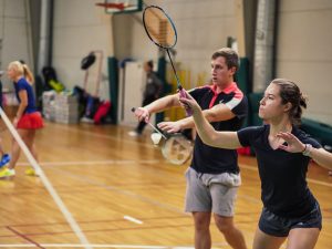 Vaikinas ir mergina žaidžia badmintoną sporto salėje. Vaikinas atlieka servavimo judesį
