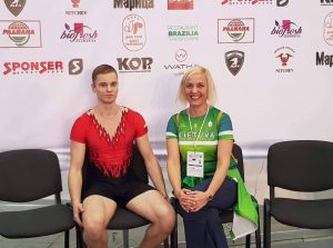Vaikinas, vilkintis raudonos ir juodos spalvų aerobinės gimnastikos triko sėdi šalia trenerės šviesiais plaukais, vilkinčios žalius marškinėlius