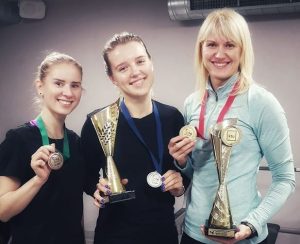 trys moterys šviesiais plaukais stovi greta ir rankose laiko medalius su apdovanojimų taurėmis