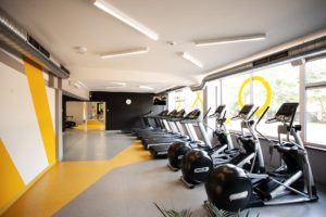 KTU ACTIVATed Gym treniruoklių salės patalpa geltonos ir pilkos spalvų sienomis bei grindimis, kurioje stovi kardiotreniruokliai
