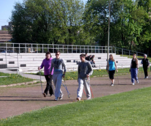 Šešios moterys su šiaurietiškomis lazdomis rankose eina stadiono bėgimo taku