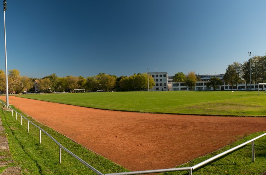 Stadionas su bėgimo taku ir žalia veja už kurio stovi keletas 4 auk&scaron;tų pastatų ir medžių.