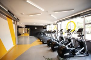 KTU Activated Gym treniruoklių salės patalpa geltonos ir pilkos spalvos sienomis ir grindimis, kurioje stovi kardio treniruokliai