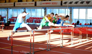 Keturi vyrai lengvosios atletikos bėgimo take atlieka šuolį per barjerus