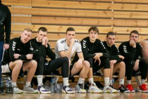 Šeši KTU krepšinio rinktinės sportininkai, dėvintys juodos spalvos aprangas, sėdi ant žaidėjų suolelio