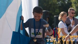 Berniukas vilkintis mėlynus marškinėlius eina rankose laikydamas apdovanojimų taurę ir mėlyną maišelį