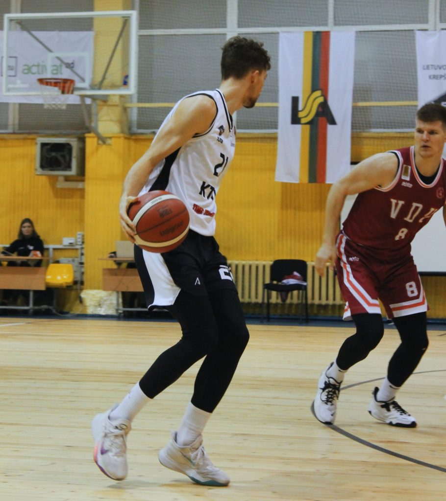 Vaikinas, vilkintis juodos ir baltos spalvos krepšinio sportinę aprangą varosi krepšinio kamuolį krepšinio aikštelėje. Greta jo bėga vaikinas tamsiai raudonos spalvos krepšinio apranga