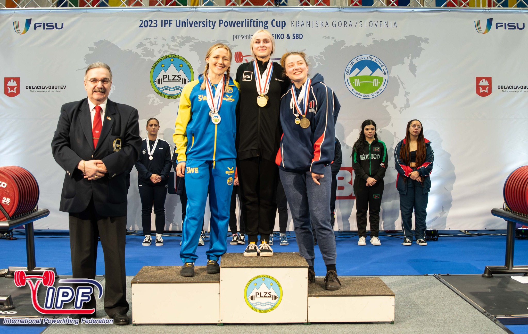 Trys merginos stovi salėje ant baltos apdovanojimų pakylos. Merginoms ant kaklo užkabinti medaliai.